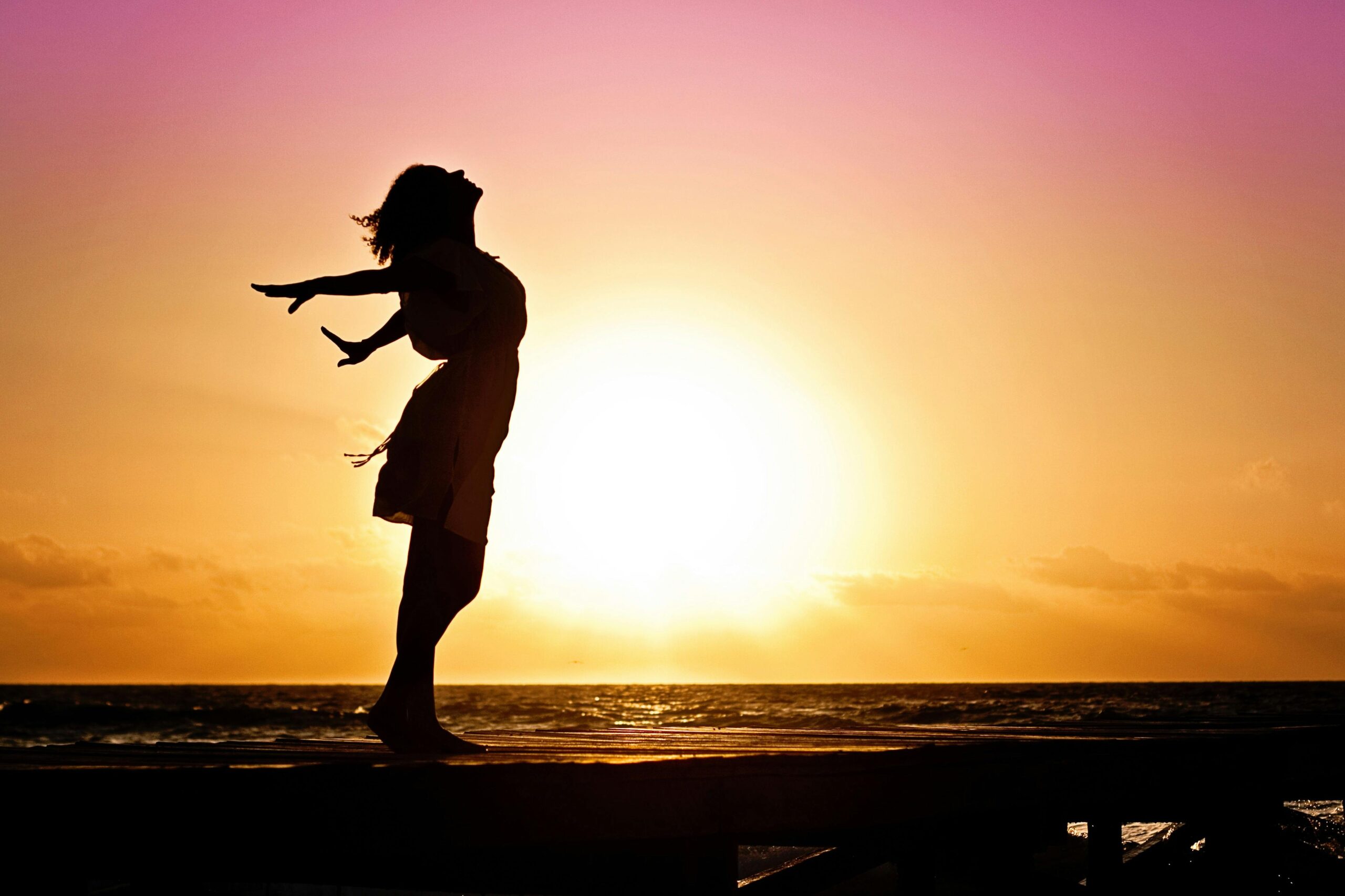 Inicio Silhouette of a woman at the beach with arms outstretched against a vibrant sunset backdrop.